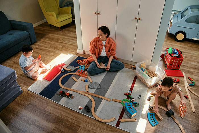 Babysitter playing with two children on floor with wooden train set as baby falls asleep in family home setting.