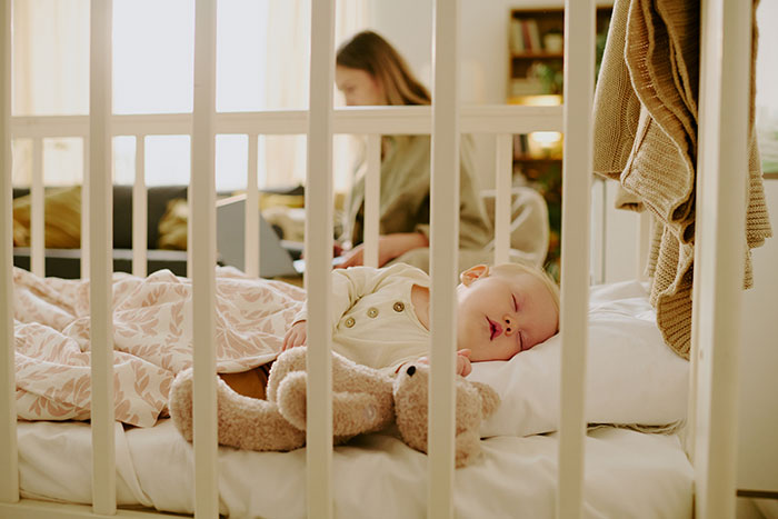 Baby falls asleep in crib with teddy bear while family member works on laptop in the background during babysitting.