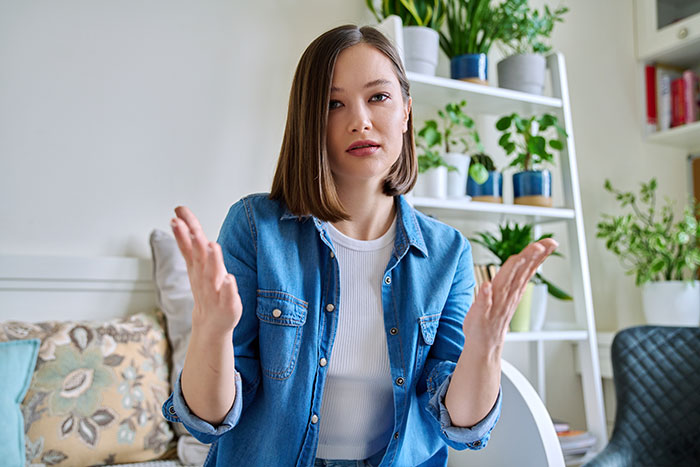 Woman in a denim shirt gesturing with hands, illustrating babysitter frustration in a home setting with plants and books.