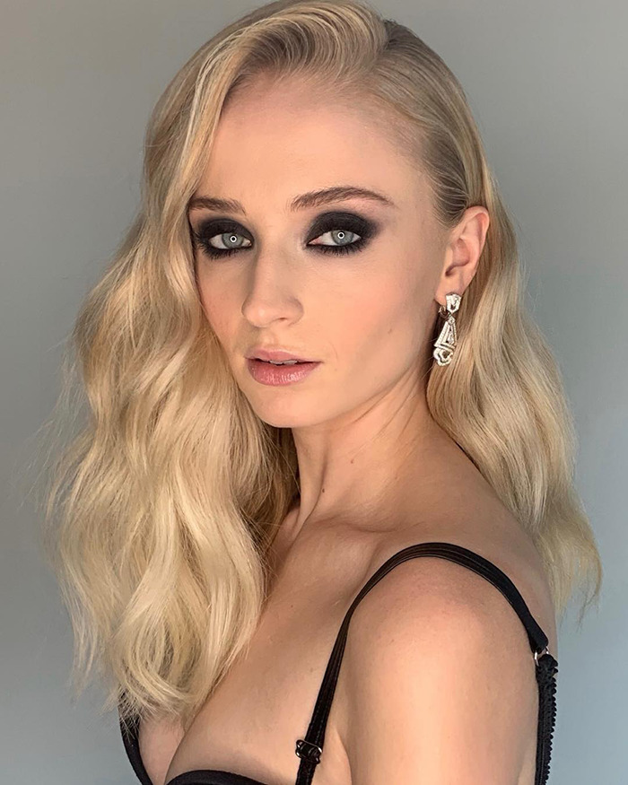 Sophie Turner posing with wavy blonde hair, smoky eye makeup, wearing elegant earrings and a black strap outfit. Sophie Turner posing with wavy blonde hair, smoky eye makeup, wearing elegant earrings and a black strap outfit.