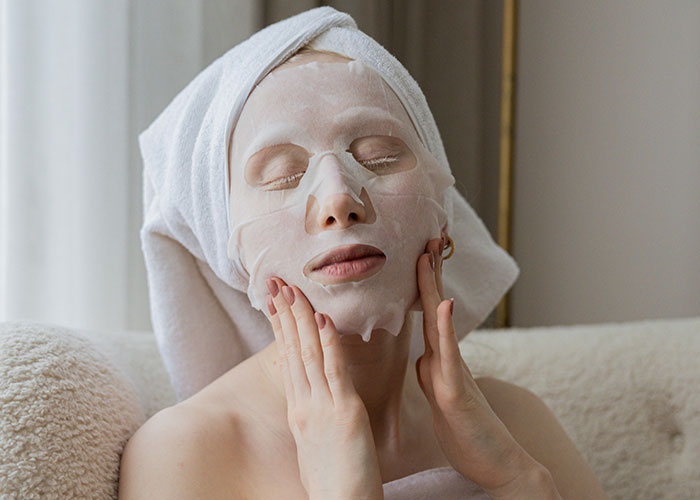 Woman relaxing with a sheet face mask and towel wrapped around her head, enjoying skincare after 30.