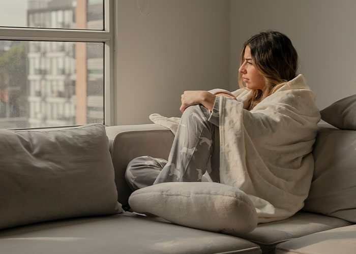 Woman wrapped in a blanket sitting on a couch near a window, reflecting quietly on things women stopped pretending they like after 30.