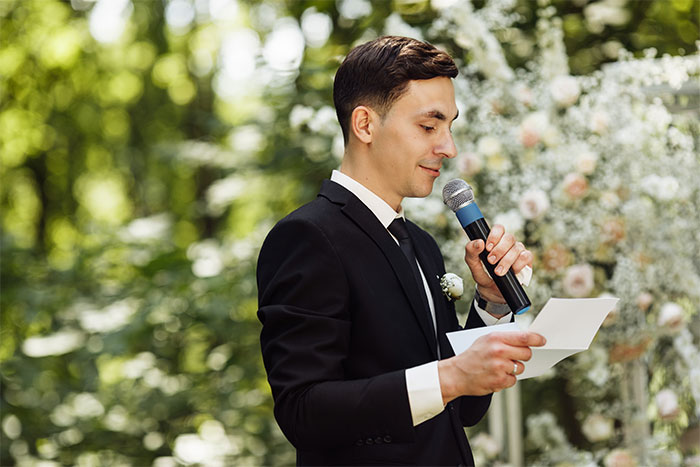 Man in a black suit holding a microphone and paper, speaking at a best friend's wedding during a proposal moment.