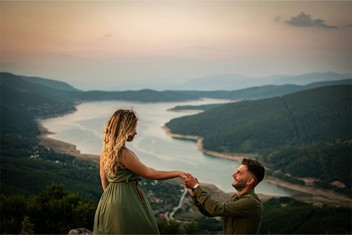 Couple overlooking a scenic river as the man kneels to propose, capturing a moment of a boyfriend&rsquo;s proposal at a wedding.