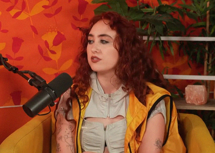 Young woman with curly red hair wearing a yellow vest, speaking into a microphone about people who knew celebrities before fame.