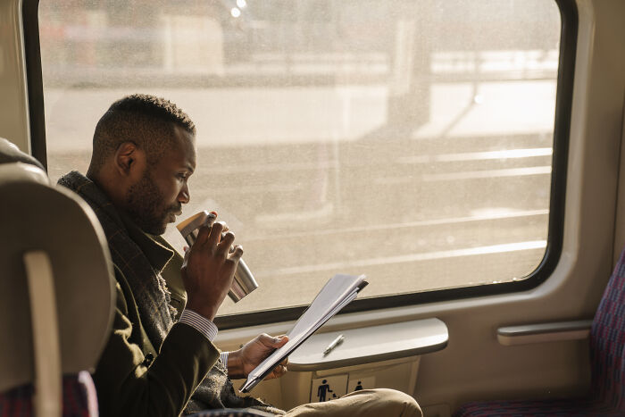 Man on a train using real-life cheat codes by reading notes and drinking coffee during daytime travel.