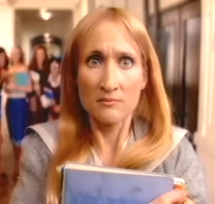 Woman with long blonde hair clutching a blue book, looking concerned in a hallway with blurred people in the background. Woman with long blonde hair clutching a blue book, looking concerned in a hallway with blurred people in the background.