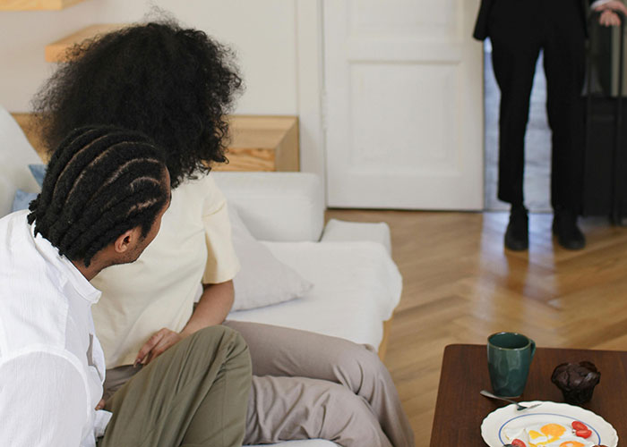 Two adults sitting on a couch looking toward a standing person, illustrating dark family secrets being revealed.