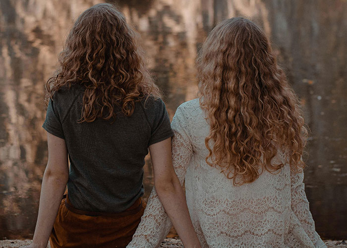 Two women with curly hair sitting by the water holding hands, reflecting on dark family secrets in adulthood.