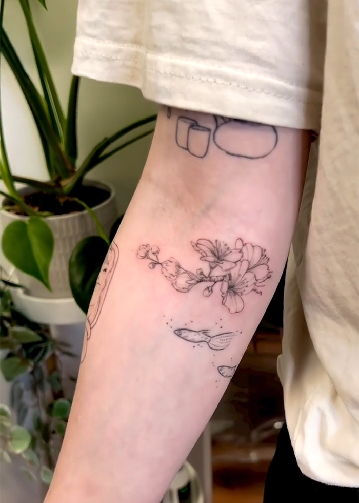 Tattoo artist drawing delicate floral and fish tattoos on a person's forearm amid indoor plants, reflecting tattoo boom trends.