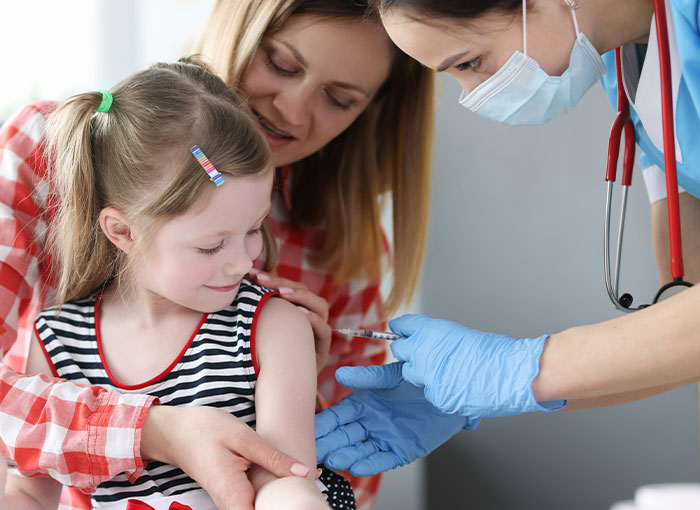 Young girl receiving vaccine from masked healthcare worker as mother looks on in medical setting related to anti-vaxxer consequences.
