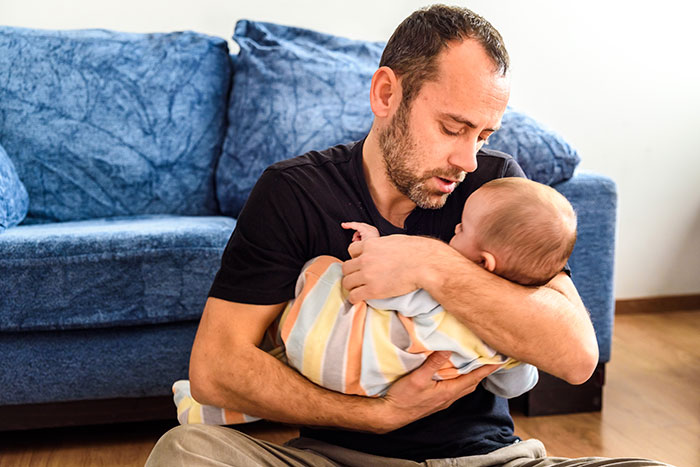 Dad sitting on floor holding baby, reflecting on feelings about joining wife's work trip with the kids at home.