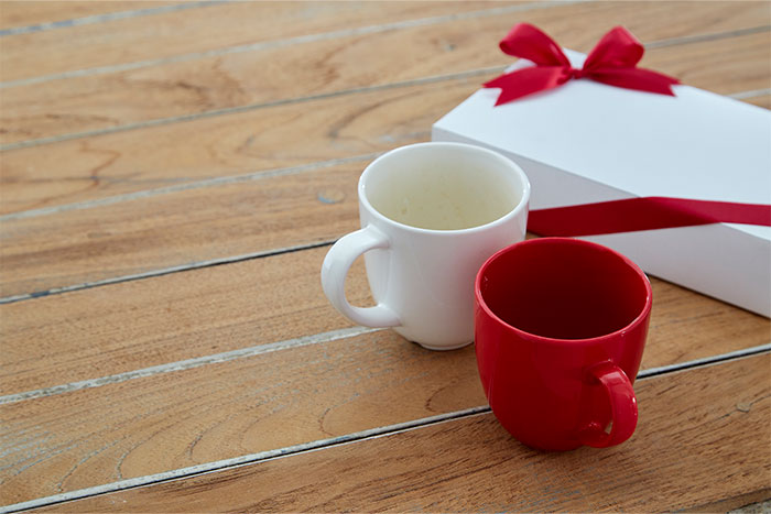 Two empty mugs, one white and one red, next to a white gift box with a red ribbon on a wooden table.