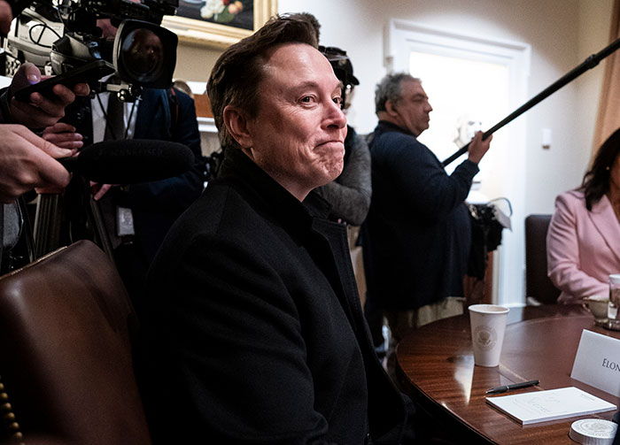 Elon Musk sitting at a table during a media event as Tesla searches for new CEO to replace him.