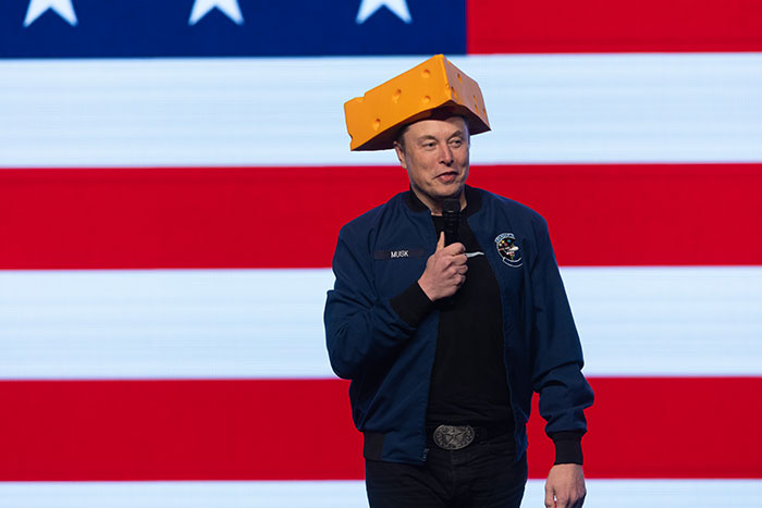 Man speaking on stage with microphone, wearing a cheese hat and jacket, against an American flag backdrop, Tesla CEO search topic.
