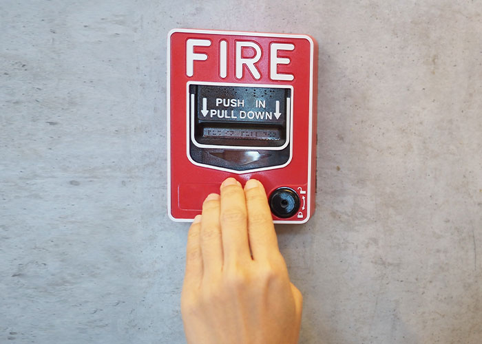 Hand reaching to activate a red fire alarm on a gray wall, highlighting emergency personnel safety equipment.