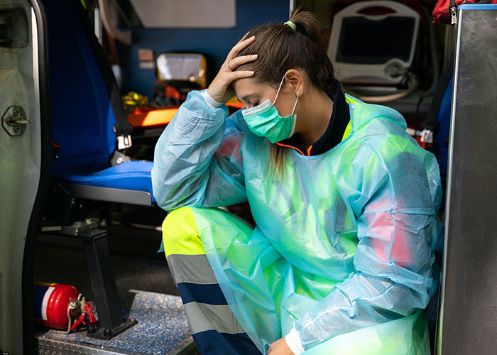 Emergency personnel in protective gear sitting inside ambulance, appearing stressed and exhausted after a shift.