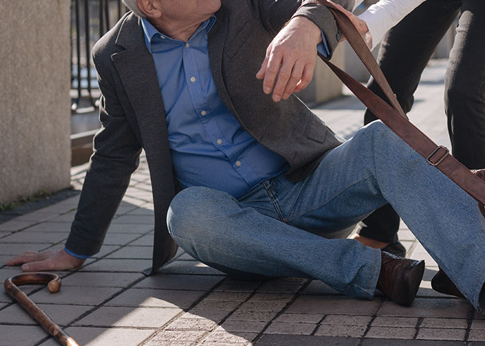 Elderly man sitting on pavement after fall while emergency personnel assist him on a city sidewalk.