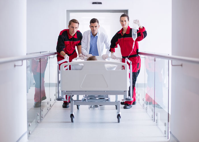 Emergency personnel in red uniforms and a doctor quickly moving a patient on a hospital bed through a hallway.