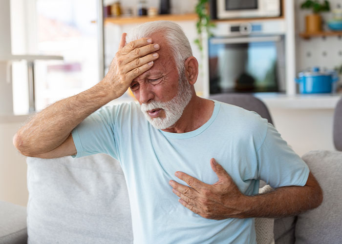 Elderly man in light blue shirt holding chest and forehead, appearing in distress, illustrating emergency personnel knowledge.