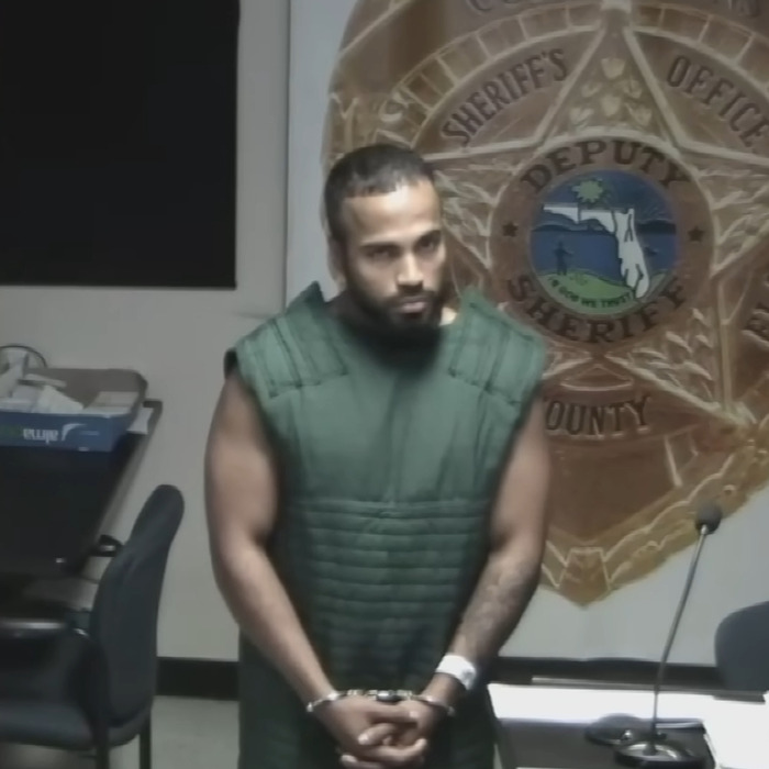 Man in green detention vest and handcuffs standing in front of sheriff's office emblem during a court appearance.