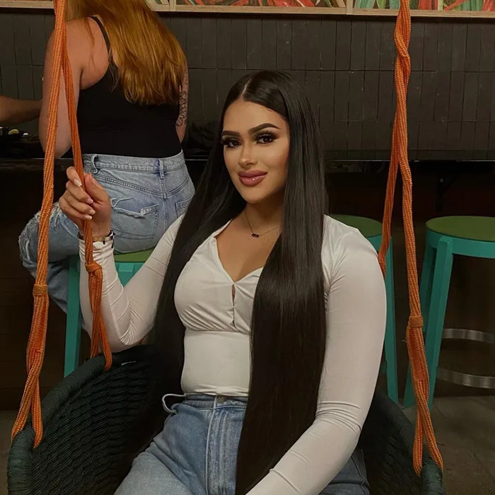 Young woman with long dark hair sitting on a swing indoors, casual outfit, smiling at the camera.
