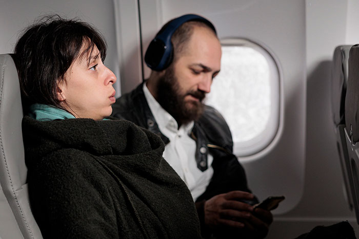 Mom defends her toddler mid-flight as angry passenger wearing headphones scrolls on phone near airplane window. Mom defends her toddler mid-flight as angry passenger wearing headphones scrolls on phone near airplane window.