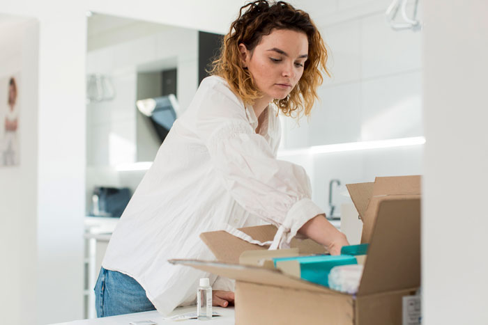 Young woman packing belongings in a box at home, reflecting on jobless partner&rsquo;s anger and toxic spending habits.