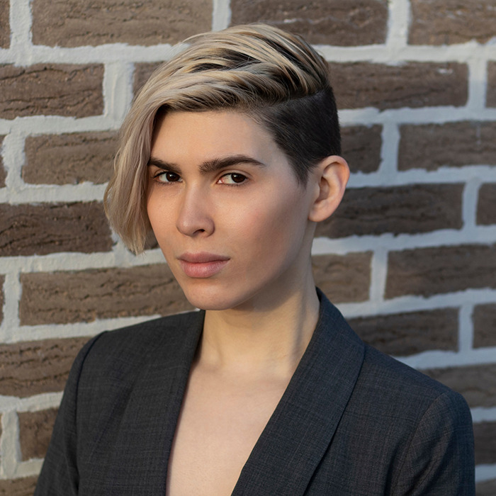 Trans archer with short blonde hair and blazer standing against brick wall, facing the camera with a neutral expression.