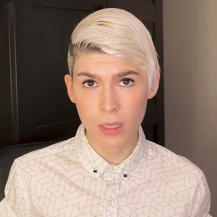 Trans archer with short blonde hair wearing a patterned shirt, speaking in an indoor setting with a serious expression.