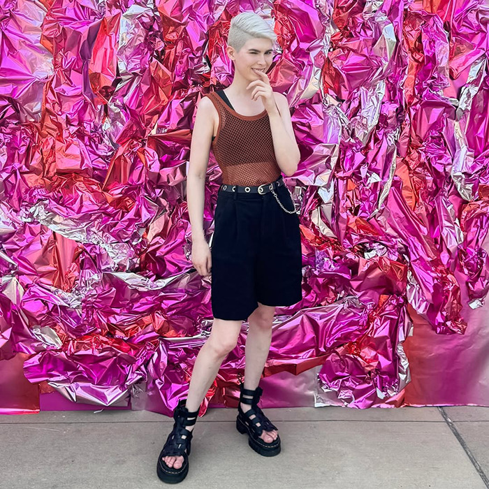 Young trans archer standing against vibrant pink metallic background, dressed in casual black shorts and a mesh top.