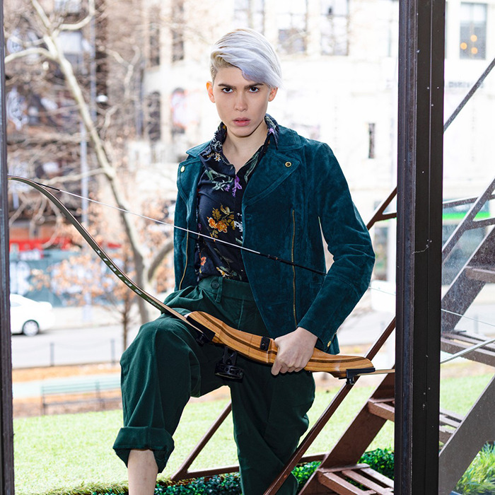 Trans archer with silver hair holding a bow outdoors, wearing a teal jacket and floral shirt, posing confidently near stairs.