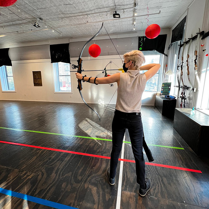 Trans archer practicing indoors aiming bow, wearing mask and light sleeveless top, with bows displayed on wall.