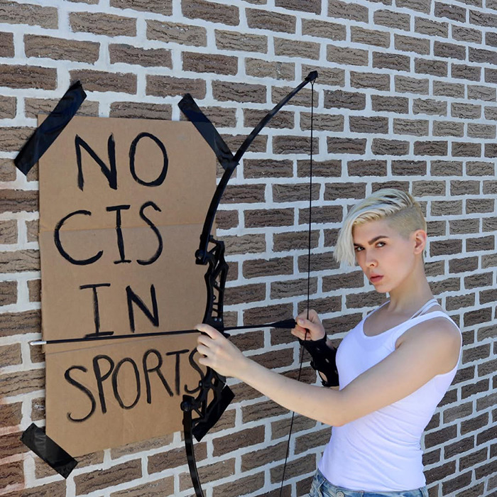 Trans archer aiming bow near wall with no cis in sports sign, illustrating backlash faced in competitive sports context.