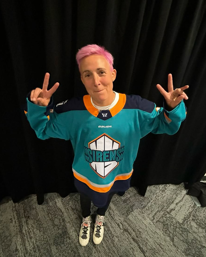 Female athlete with pink hair wearing teal and orange hockey jersey posing with peace signs against black curtain.