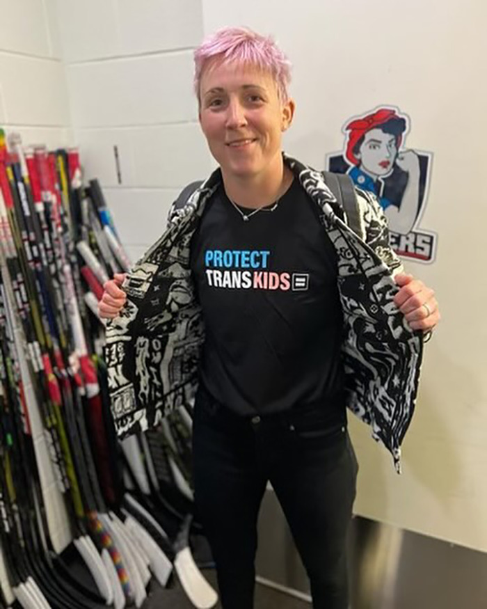 Female athlete showing support for trans kids wearing jacket open in a hockey locker room with sticks in background