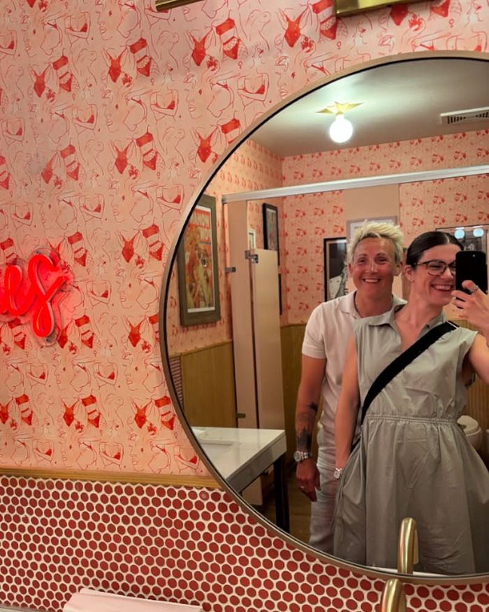 Two women smiling and taking a selfie in a women's restroom with retro decor and patterned wallpaper.