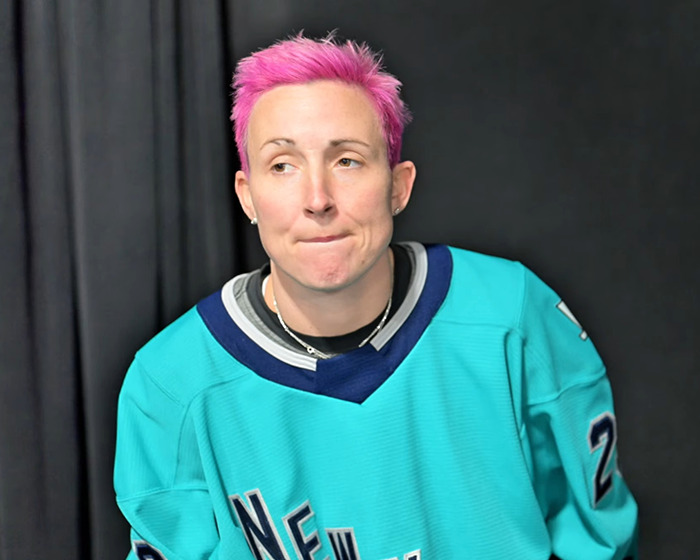Female athlete with short pink hair in teal sports jersey, looking away with a thoughtful expression against a dark background.