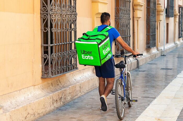 Delivery person with Uber Eats bag walking a bicycle along a street, representing people share companies bad list concept.