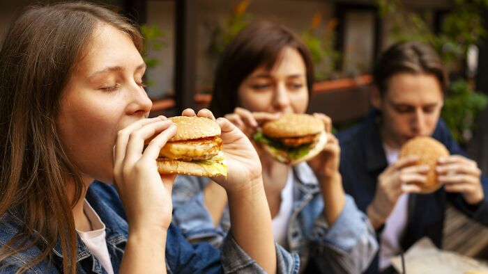 Three young women eating unhealthy fast food burgers outdoors showcasing unhealthy eating habits observed in others.