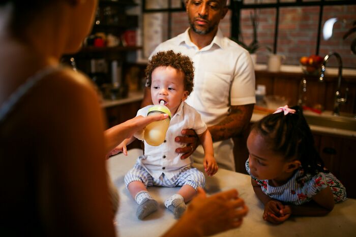 Family scene showing a toddler drinking from a bottle while adults supervise unhealthy behaviors observed by others.