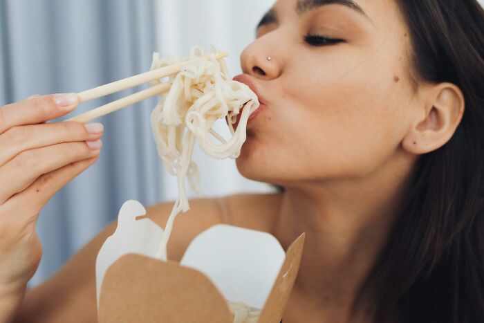Young woman eating noodles with chopsticks from a takeaway box, illustrating unhealthy human things related to diet.