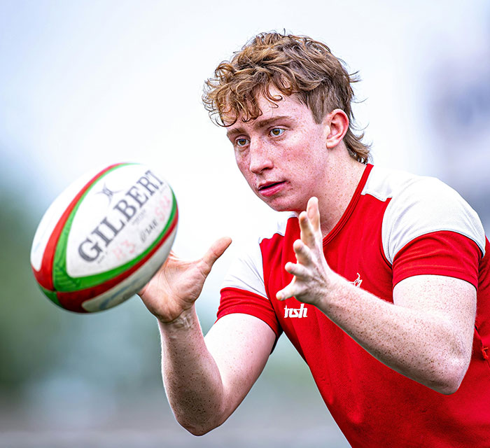 Teenage boy focused on catching a rugby ball wearing a red and white sports shirt outdoors.