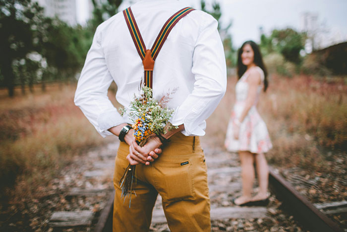 Man holding flowers behind his back wearing suspenders, woman standing on railway tracks in floral dress, wedding theme.