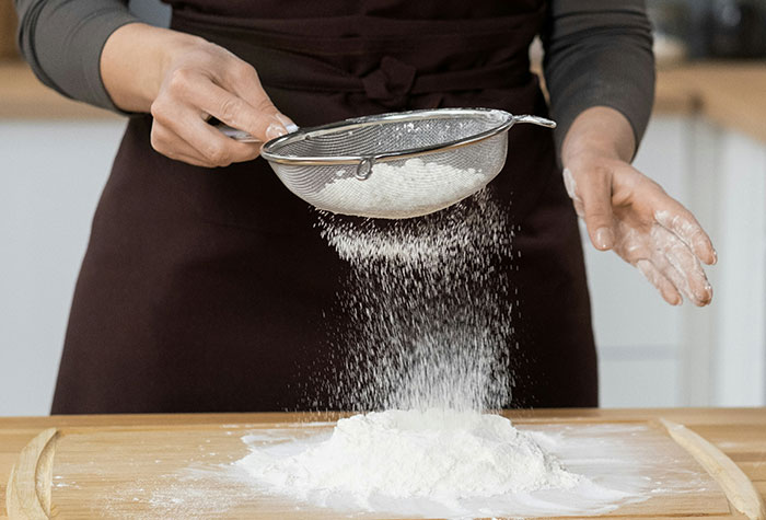 Person sifting flour through a metal sieve over a wooden surface, demonstrating common cooking advice in a kitchen.
