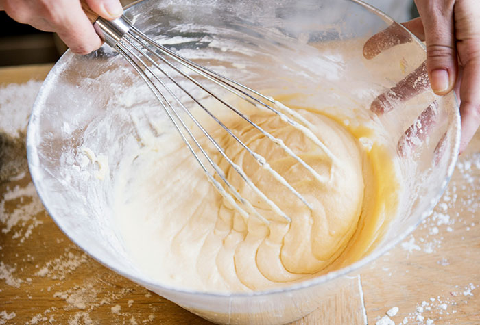 Hands whisking batter in a glass bowl with flour on the wooden surface, illustrating common cooking advice tips.