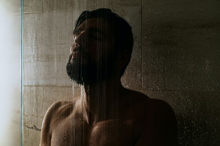 Man enjoying low-key pleasures standing under warm shower water in a dimly lit bathroom setting.