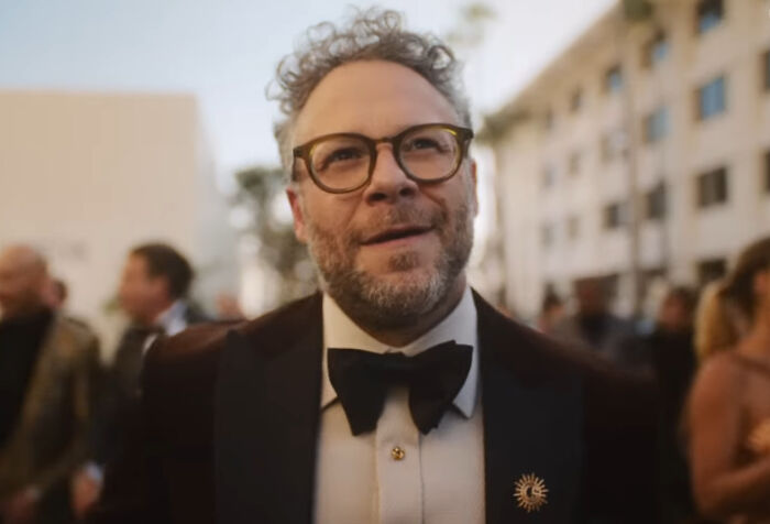 Man in glasses and bow tie sharing stories from behind the tap at a lively bartenders event outdoors.