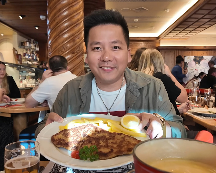 Vlogger smiling while holding a plate of food at a restaurant unaware he is sitting next to an A-list star nearby.
