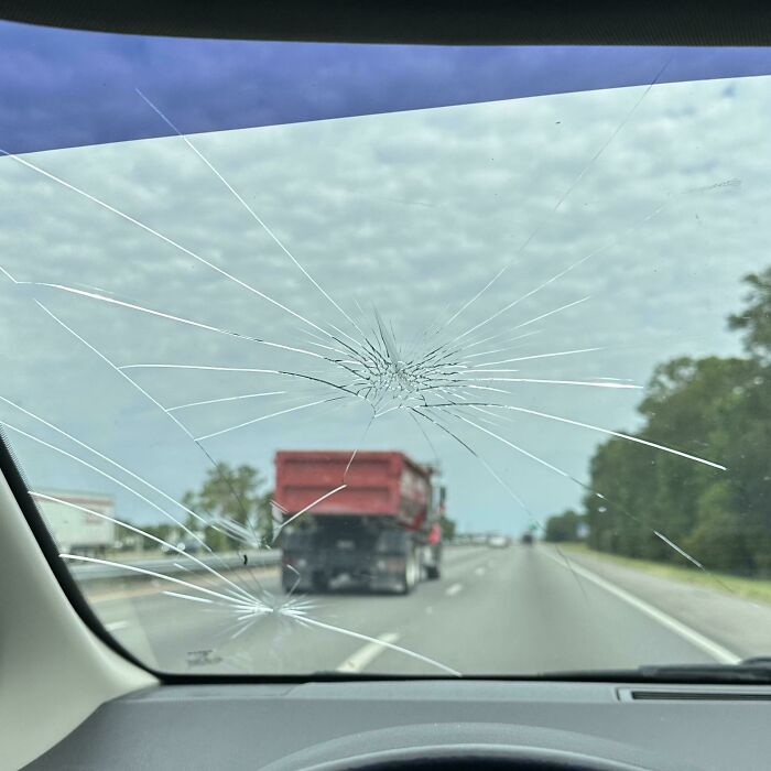 View from inside a car with a c*****d windshield looking at a red truck on a highway during the day.